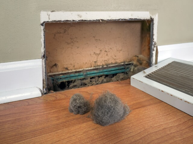 A neglected air vent filled with dust and two large dust bunnies on the floor highlights the importance of home maintenance.